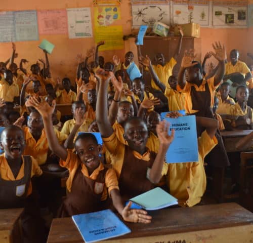 School children in a classroom sitting at desks with their hands in the air holding SPANA Humane Education work books