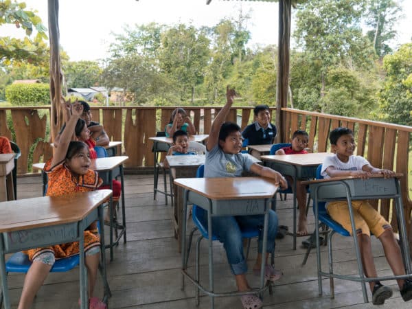 School children in Costa Rica sitting outside at desks with their hands up to answer a question