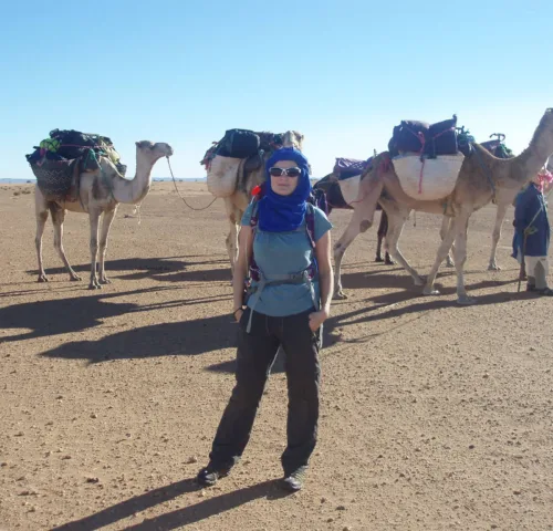 A lady stands in front of three camels in the Sahara Desert as she participates in a trek to raise funds for SPANA.