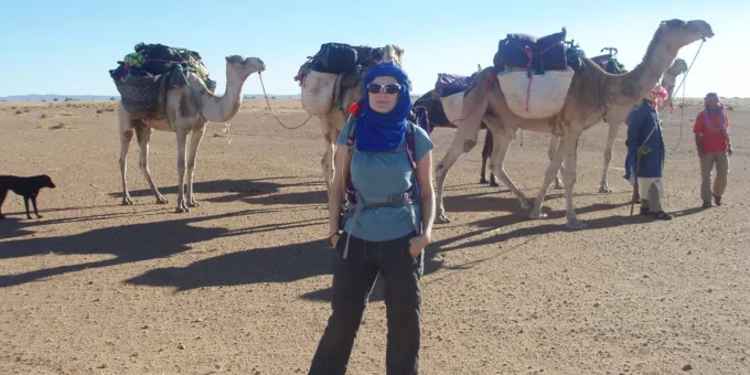 A lady stands in front of three camels in the Sahara Desert as she participates in a trek to raise funds for SPANA.