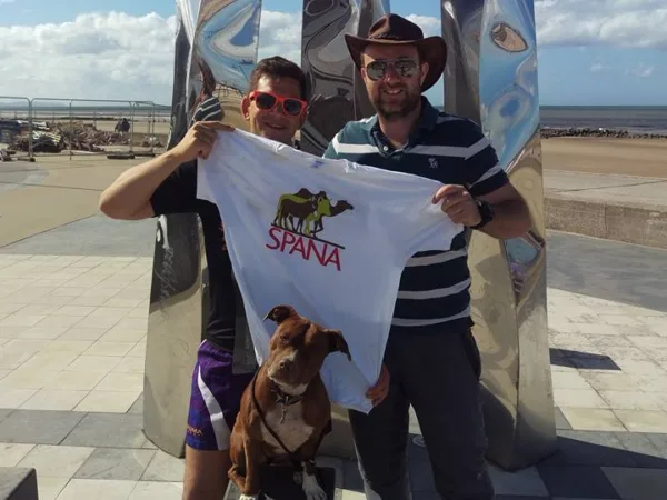 Two men are smiling as they pose with a dog and a SPANA shirt.