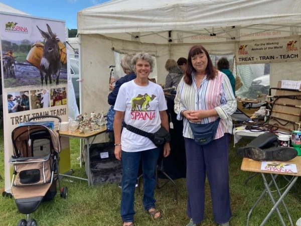 Two ladies standing in front of a SPANA kiosk and SPANA sign raise money for working animals at a fundraising event.