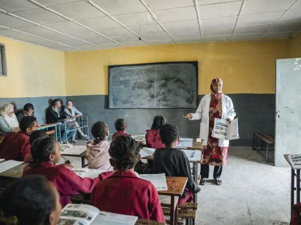 a group of school children sit at their desks facing away from the camera and a teacher stands at the front of the classroom. Behind her is a blackboard