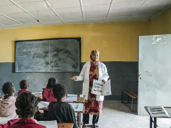 a group of school children sit at their desks facing away from the camera and a teacher stands at the front of the classroom. Behind her is a blackboard