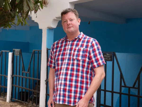 A photo of Tim Coggan wearing a blue, white and red checked shirt, standing in front of stables that are painted blue with black gates.